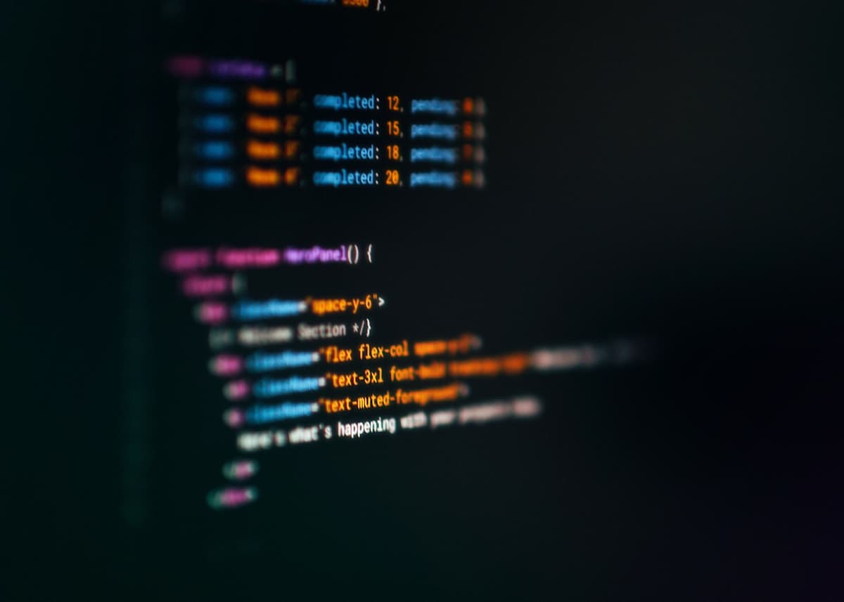 Layar laptop menampilkan kode JSX dengan syntax highlighting warna-warni di ruangan gelap.