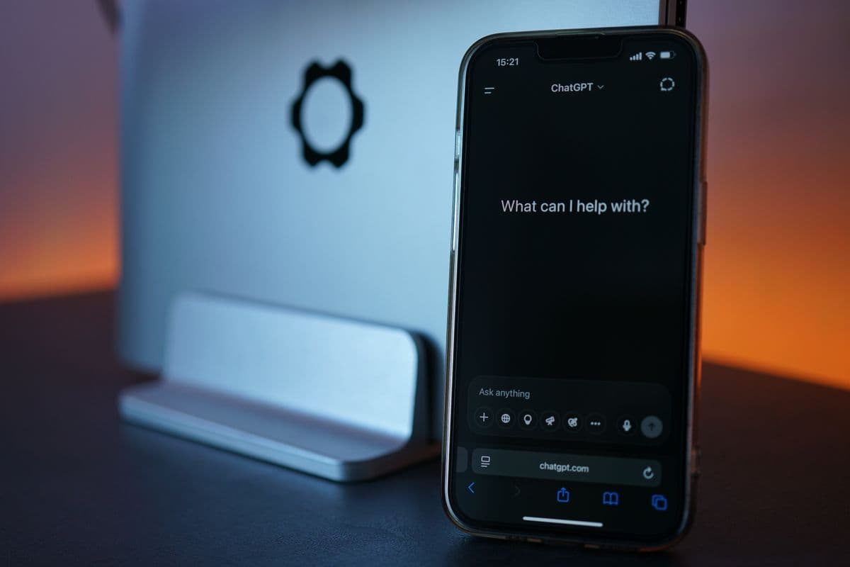 Layar smartphone menampilkan aplikasi ChatGPT dengan teks 'What can I help with?', dengan laptop di latar belakang bercahaya oranye.