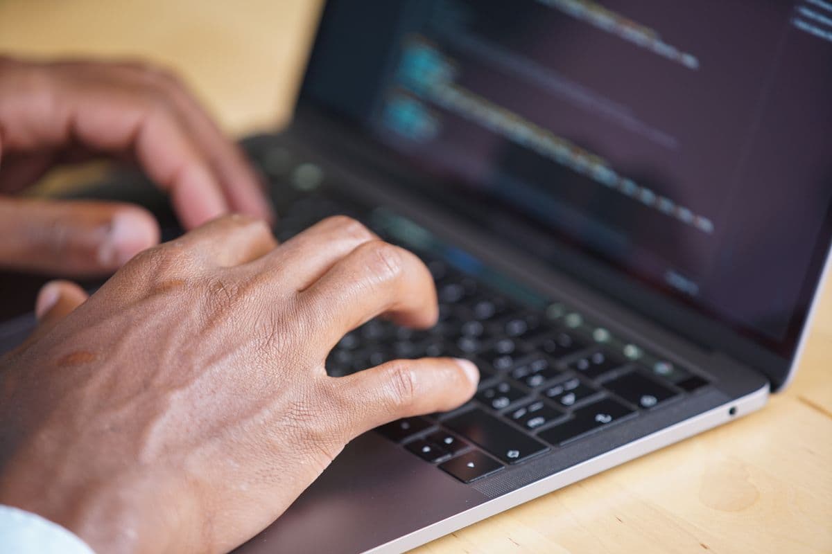 Tangan mengetik di keyboard laptop dengan layar menampilkan teks kode bertema gelap.