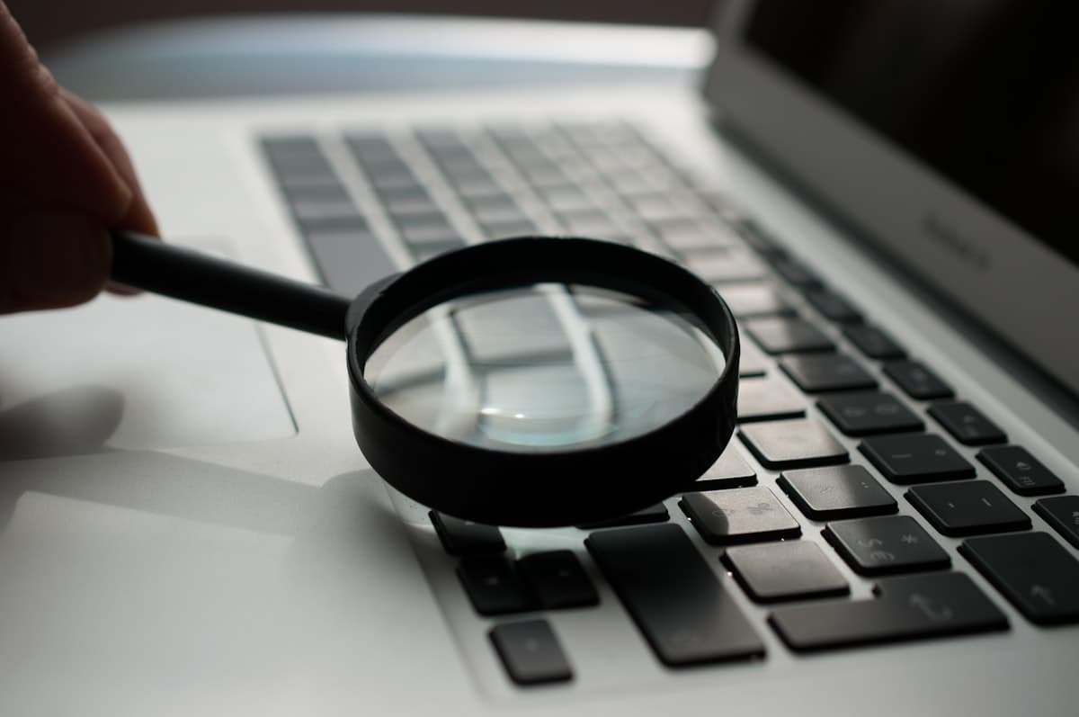 Kaca pembesar diletakkan di dekat laptop berwarna abu-abu di atas meja kerja, menggambarkan aktivitas pencarian dan riset.