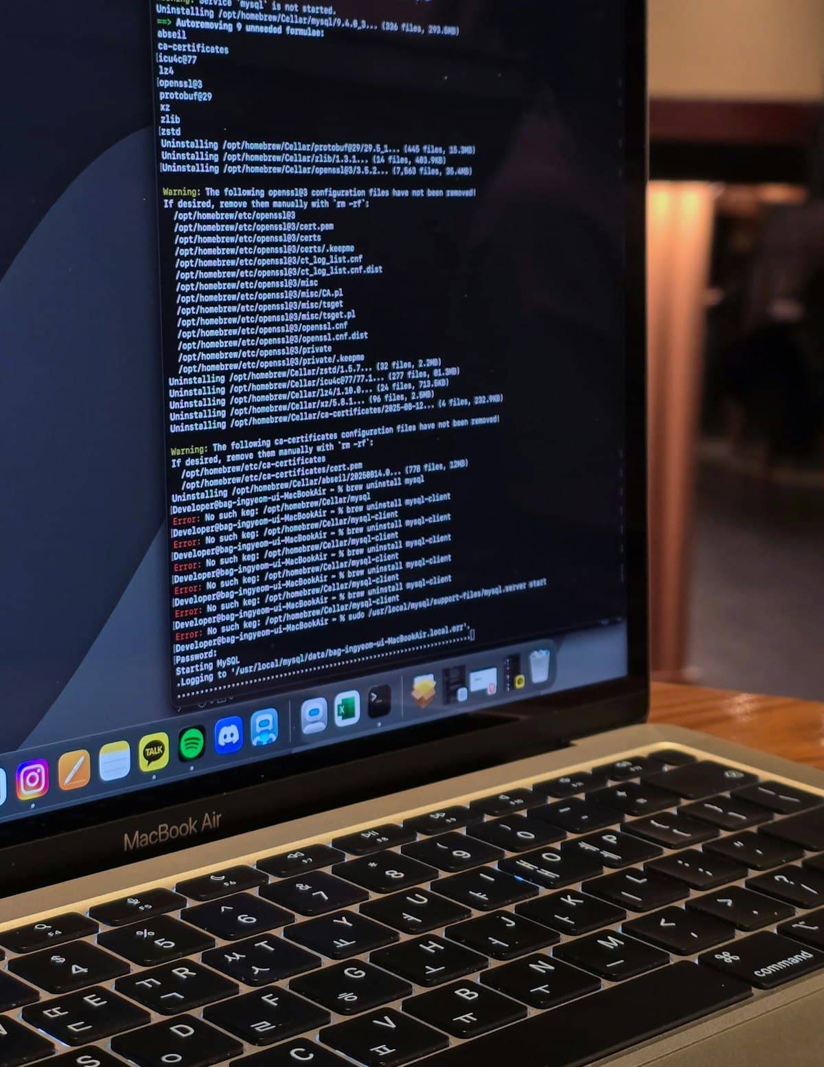 MacBook menampilkan baris perintah terminal dan log kode di ruang kerja dengan pencahayaan hangat.