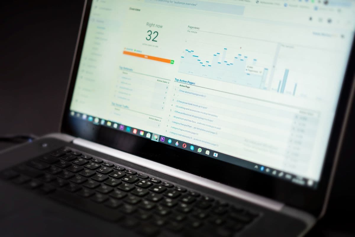 Laptop dari sudut miring menampilkan dasbor analitik web dengan grafik pageview per menit dan daftar halaman paling aktif di atas meja gelap.